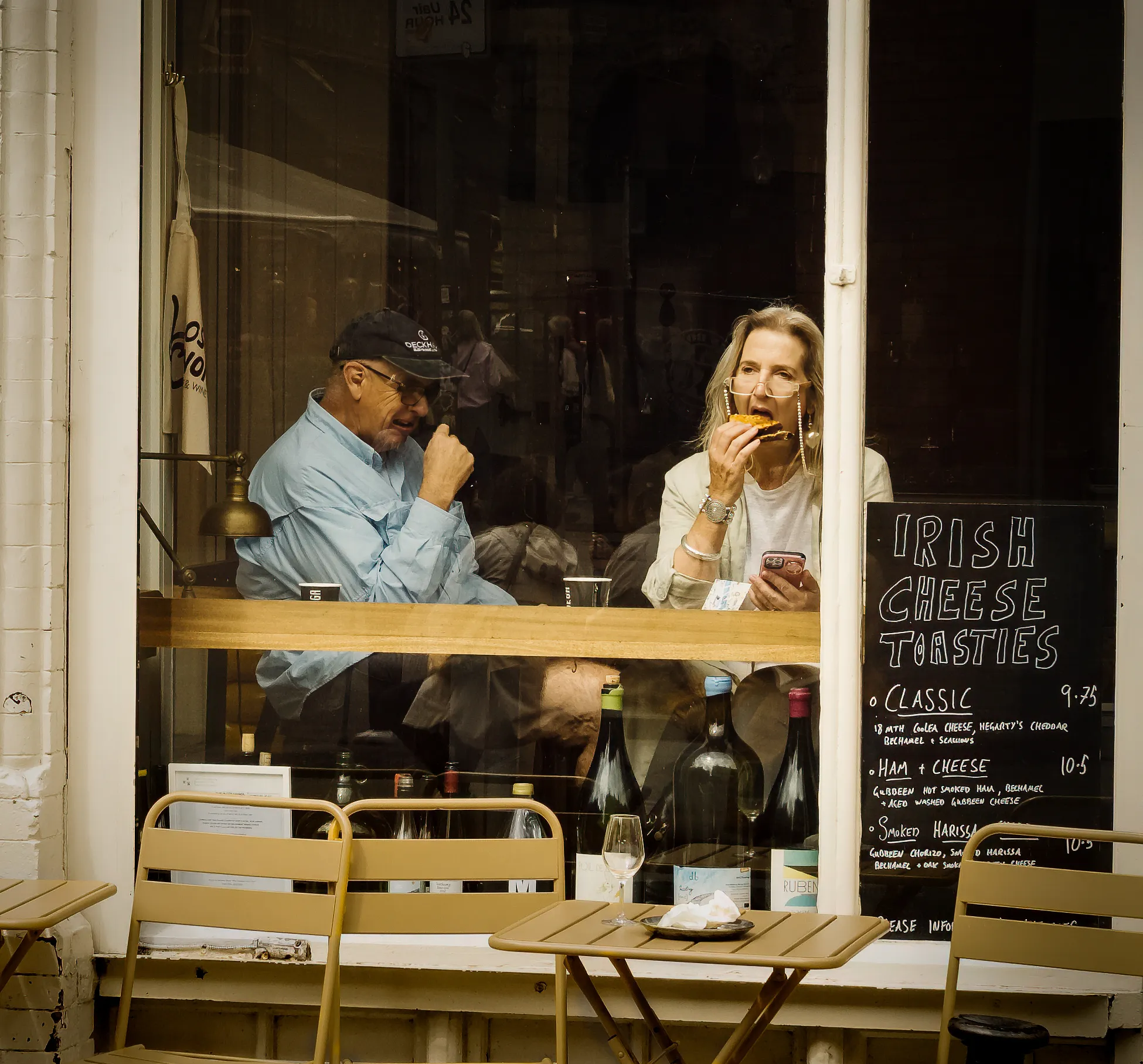 A woman eats a sandwich in a deli window while a man looks on