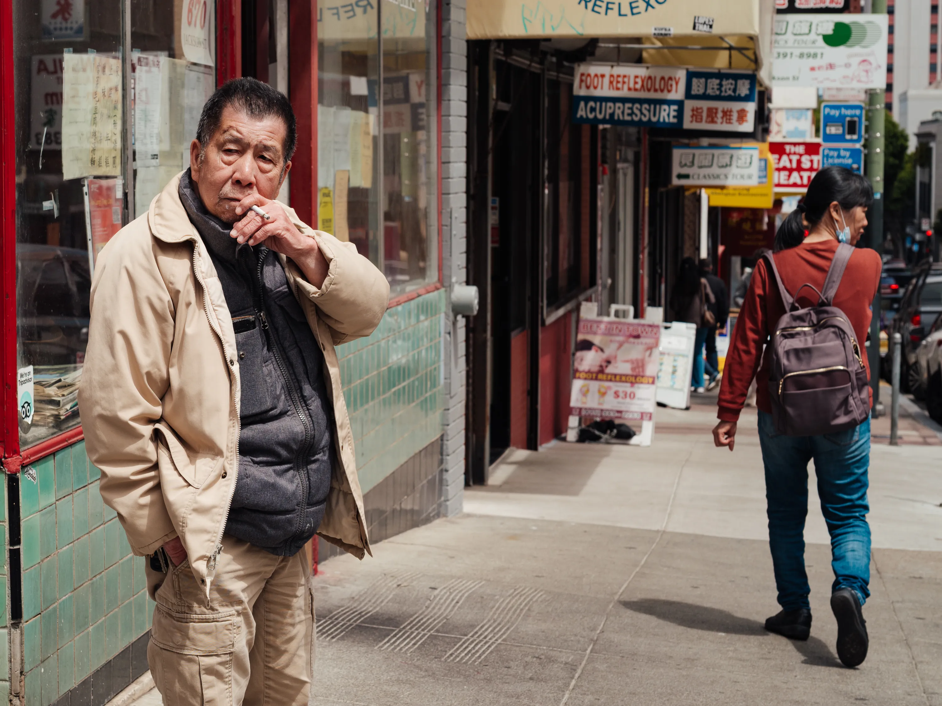 A man smokes a cigarette while a woman walks down the sidewalk in a mask